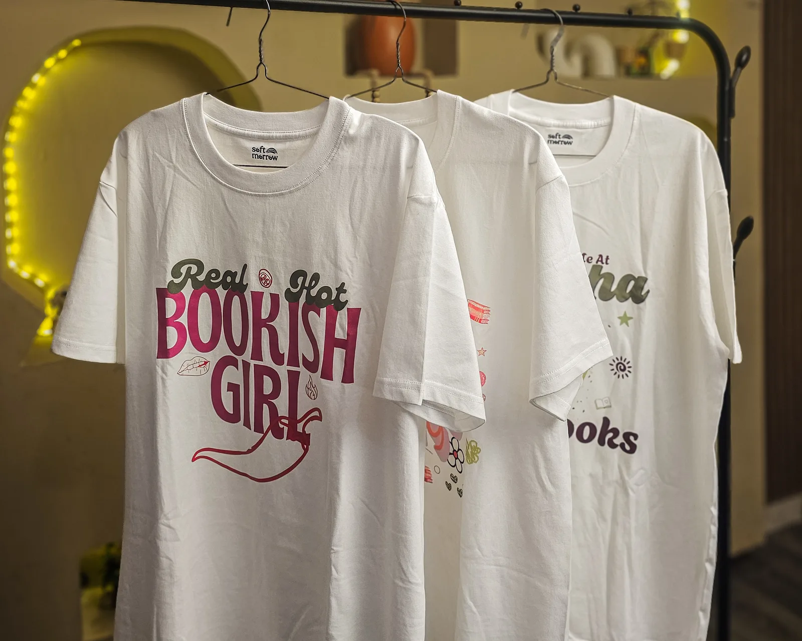 A rack of white Softmorrow tees, including the Real Hot Bookish Girl tee, against a warm sculpted wall with soft lighting